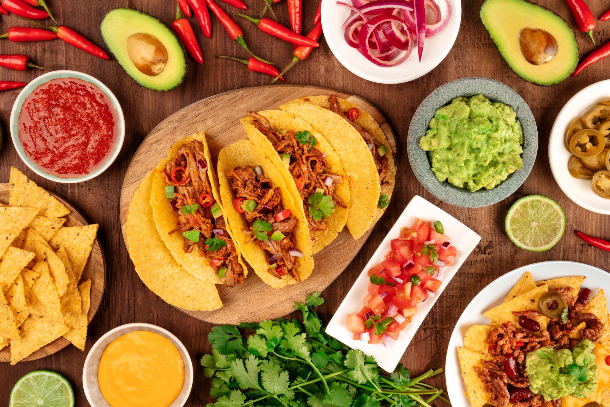 A photo of Mexican food, including tacos, guacamole, pico de gallo, nachos and others, shot from the top with ingredients on a dark rustic wooden background